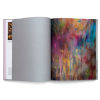 An open art book on a white surface. The right page displays a full-bleed, vibrant abstract painting with streaks and scribbles of magenta, blue, and yellow. The left page is mostly covered by a blank, light gray flyleaf.