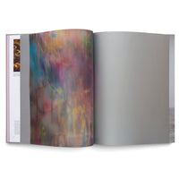 An open art book seen from an angle on a white background. The left-hand page shows a full-bleed, vibrant abstract image with blurred vertical streaks of pink, blue, and yellow. The right-hand page is a solid, blank, light grey sheet.