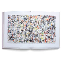 An open book on a white surface shows a two-page spread of a large, complex abstract artwork. The piece is a dense composition of black, graffiti-like scribbles and energetic lines over a background with splashes of color, including orange, yellow, red, and purple. The artwork is visually divided into a grid of twelve rectangular panels.