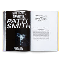 An overhead shot of an open book with a yellow cover. The left page has a black background with bold, white German text reading Sauerteigbrot, Alpenbutter, Patti Smith, and a black-and-white photo of a dog with glowing eyes. The right page is white and features the beginning of an interview in German with Hans Ulrich Obrist.