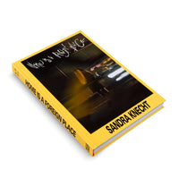 A photograph of a hardcover book with a bright yellow cover and spine, resting on a white surface. The book is titled Home Is A Foreign Place by Sandra Knecht in bold black text. The cover features a dark, blurry photo of a person with long blonde hair on a city street at night, with motion-blurred lights in the background. The title is also written in a white, graffiti-style script over the image.