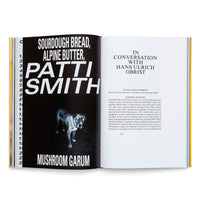 An open book with a yellow spine showing a two-page spread. The left page has a black background with large white text reading Sourdough Bread, Alpine Butter, Patti Smith, and Mushroom Garum, along with a black-and-white photo of a dog. The right page is white with black text, featuring the heading In Conversation with Hans Ulrich Obrist above an interview transcript.