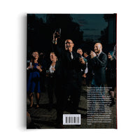 The back cover of a book, showing a photograph of a celebratory outdoor gathering at night. A man in a suit and glasses stands in the center, laughing and raising a glass in a toast, surrounded by smiling guests. Below the image is a block of German text, a website address, and a barcode.
