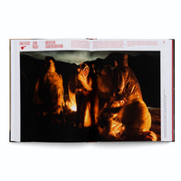 A photograph by John Moore displayed in an open book. The image captures a group of migrants at night, wearing translucent plastic ponchos and huddled together on rocky terrain. A warm, orange light, likely from a fire, illuminates them from a low angle, creating a stark contrast with the surrounding darkness.
