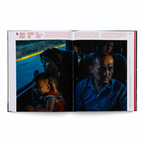 An open book with a photo spread across two pages, showing portraits of weary travelers on a bus. The left page shows a woman holding a young child, both illuminated by the cool, blue light of dusk from the window. The right page is a close-up of a man's face, half-lit, with a somber and exhausted expression. German text frames the photographs.