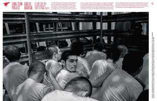 A wide, black and white photograph by Carlos Barrera captures a large, dense crowd of incarcerated men with shaved heads in El Salvador. They are packed tightly together in a holding cell, most with their backs to the viewer. In the center, one man turns his head to look directly at the camera with a somber expression.