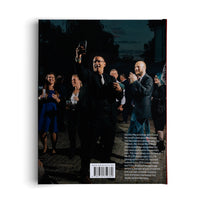 The back cover of a book, featuring a nighttime photograph of a joyous man in a black suit and glasses raising his glass in a toast. He is surrounded by a smiling crowd at an outdoor gathering. Below the image is a text description of the book, a website address, and a barcode.