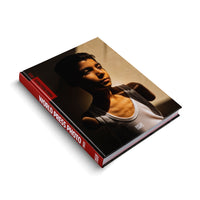 A black hardcover book, World Press Photo 2025, is displayed at an angle on a white background. The red spine has white text. The front cover features a striking photograph of a young person in a white tank top, their face and chest partially illuminated by a strong, diagonal beam of light against a shadowy backdrop.