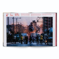 An open book displays a two-page photograph with the headline GAZA UNDER ISRAELI ATTACK. The photo shows a street scene at dusk where a building in the background is exploding in a cloud of dark smoke and a shower of fiery sparks. In the foreground, people are silhouetted as they stand on the street and watch the destruction.