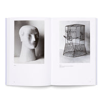 An open book on a white surface showing two black-and-white photographs of head sculptures. The sculpture on the left is a solid, white, stylized bust. The sculpture on the right is a contrasting wireframe, cage-like construction.