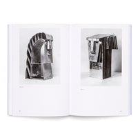 An open art book lies flat on a white surface, displaying a two-page spread. Each page features a black-and-white photograph of an abstract, cubist-style metal sculpture of a head against a plain backdrop. The page numbers 98 and 99 are visible at the bottom.