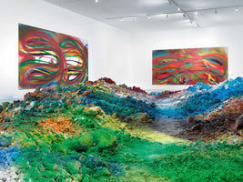 An art gallery installation featuring a floor covered in large, colorful mounds of powdered pigment in greens, blues, yellows, and oranges, with a path winding through the center. On the white gallery walls in the background hang two large, abstract paintings with vibrant, swirling colors that echo the installation on the floor.