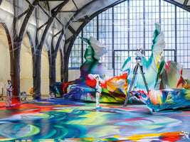 Two artists in white hazmat suits create a massive, colorful abstract art installation in a large hall with high, arched ceilings. The entire floor and several towering sculptures are covered in vibrant, swirling streams of red, blue, and yellow paint. One artist stands on a ladder, spray-painting a sculpture, while the hall's columns are wrapped in protective plastic.