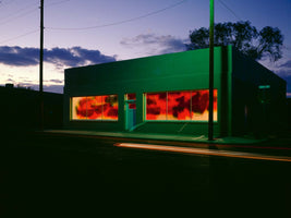 A long-exposure photograph of a one-story building at dusk, located on a street corner. The building's facade is bathed in a vivid green light, while its large windows are illuminated from within, revealing a fiery, abstract pattern of red, orange, and black. In the dark foreground, red and white light trails from a passing vehicle streak across the pavement.