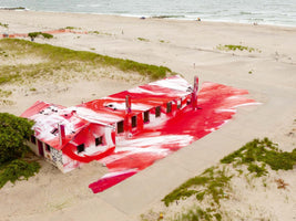 An aerial view of a long, derelict building on a sandy beach, covered in a massive, swirling art piece of red and white paint that spills from the roof onto the surrounding pavement, with the ocean in the background.