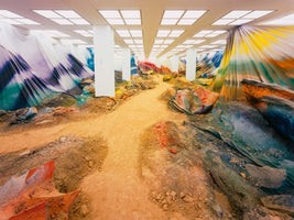 An expansive art installation inside a gallery with white pillars and a grid of ceiling lights. A winding dirt path cuts through a fabricated landscape of soil and rocks. The walls are draped in massive, flowing fabrics painted with vibrant blends of yellow, orange, blue, and purple.