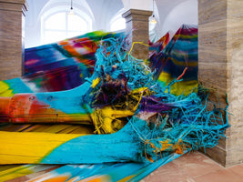 A vibrant and chaotic art installation in a gallery featuring large, uprooted tree trunks and tangled roots spray-painted in bold shades of blue, yellow, orange, and purple. The sculpture is set against a matching multicolored fabric backdrop between stone pillars.