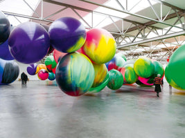 An immersive art installation in a large, high-ceilinged gallery where dozens of giant, multicolored spheres float at various heights above a polished concrete floor. A few people wander among the massive orbs, which are painted in vibrant shades of purple, green, yellow, and pink, showcasing the immense scale of the artwork.