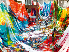 An overhead view of a massive, immersive art installation inside a large warehouse. People walk through a valley created by enormous sheets of brightly colored, tie-dye style fabric that hangs from the ceiling and pillars, covering the floor in vibrant waves of red, blue, orange, and yellow.