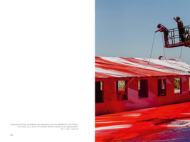 A two-page book spread. The right page shows two people on a mechanical lift spray-painting a single-story building and the ground with vibrant red and white paint under a clear blue sky. The roof is painted with red and white stripes, and the ground is saturated with red. The left page contains a caption in German.