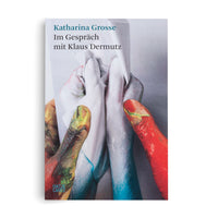 The cover of the book Katharina Grosse Im Gespräch mit Klaus Dermutz. The cover art is a close-up photograph of hands covered in thick, wet paint. Fingers smeared with red, yellow, and green interact with other fingers coated in glossy white and blue paint against a pale background.