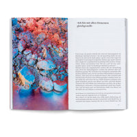 An open book viewed from above on a white surface. The left page shows a full-bleed close-up photo of a textured, crumbly substance in vibrant red, orange, and blue. The right page contains German text with a large heading, Ich bin mit allen Elementen gleichgestellt, and several paragraphs below it, with the page number 45 in the corner.