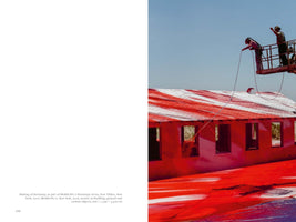 A two-page book spread. The right page features a photograph of two people on a cherry picker spray-painting a single-story building and the ground below with vibrant red and white paint under a clear blue sky. The building's roof is covered in wide red and white stripes, and the ground is a sea of bright red. The left page contains text describing the art installation.