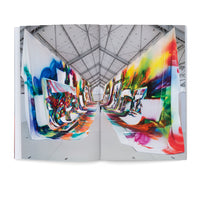 An open book displays a two-page photograph of a monumental art installation within a vast, high-ceilinged industrial hall. Two continuous, massive banners covered in vibrant, abstract, multi-colored paintings are draped from the ceiling on either side of a central walkway. The fabric creates a flowing, canyon-like path, and a tiny human figure walking in the distance emphasizes the artwork's immense scale.