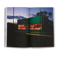 An open book displays a grainy, two-page photograph of a street corner at dusk. A modern, dark green building is illuminated by its large windows, which glow with a warm, orange-red light, backlighting graffiti on the glass. In the foreground, a streak of light from a passing car cuts across the dark road under a twilight sky.