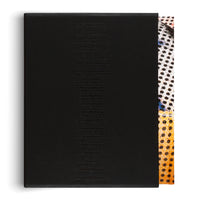 A matte black slipcase with a list of names embossed vertically in black text, shot against a white background. A book with a colorful cover featuring black polka dots on white and yellow-orange sections is partially visible peeking out from the right side of the case.
