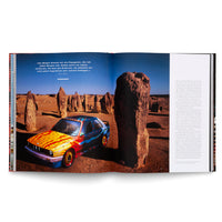 An open book showing a two-page photo spread of a brightly painted BMW Art Car parked in a desert landscape. The car is covered in colorful yellow, red, and blue abstract patterns and is surrounded by tall, weathered rock formations under a clear blue sky.