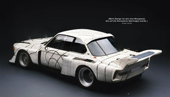 A studio photograph of the BMW 3.0 CSL Art Car designed by Frank Stella, shown from the rear three-quarter view against a dark background. The entire white body of the race car is covered in a black and white grid pattern resembling graph paper, with bold, black, geometric and curving lines overlaid on top, creating the impression of a blueprint.