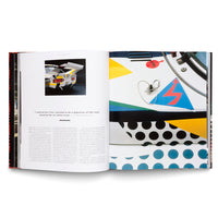 An open book on a white surface shows a two-page spread about the BMW Art Car by Roy Lichtenstein. The left page has text, a quote, and a photo of the car's rear. The right page is a large close-up of the car's hood, featuring colorful pop art graphics like polka dots, geometric shapes, and a red lightning bolt.