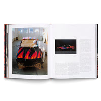 An overhead shot of an open book with a red spine, laid flat on a white surface. The left page features a large photo of an artist in a white suit and mask painting vibrant red, orange, and blue flames onto the hood of a black car in a workshop. The right page shows a smaller photo of the completed car and two columns of text.