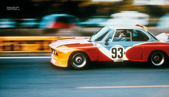 A panning shot of the Alexander Calder BMW Art Car, a 3.0 CSL with the number 93, racing at the 24 Hours of Le Mans in 1975. The car is painted in bold blocks of red, yellow, and blue, and is captured in motion, with the track and background blurred to convey a sense of speed.