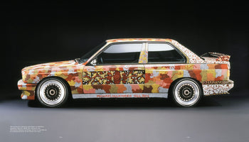 A side-profile studio shot of the 1989 BMW M3 Art Car by Michael Jagamara Nelson. The entire vehicle is covered in an intricate Australian Aboriginal Papunya Tula dot painting. The artwork uses a warm, earthy palette of red, orange, yellow, and grey to form complex geometric patterns and symbols across the car's body, set against a dark background.