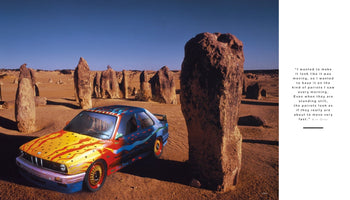 A brightly colored BMW Art Car by Ken Done sits parked in a sun-drenched desert. The car's hood is yellow with spots, and its sides feature flowing red and blue abstract patterns. The vehicle is surrounded by tall, weathered limestone formations in the Pinnacles Desert under a clear, deep blue sky.