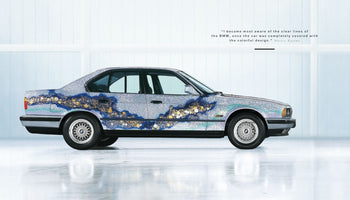 A side profile of the BMW Art Car by Matazo Kayama, a 5-Series sedan, in a brightly lit studio. The car's body is covered in a silver foil-like texture, with a wide, flowing band of dark blue, gold, and silver leaf patterns running along its side, resembling a traditional Japanese design.