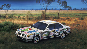 A BMW sedan art car painted with vibrant, colorful Ndebele geometric patterns sits in a grassy savanna. The car's intricate design, featuring pink, blue, yellow, and green shapes outlined in white, contrasts with the natural landscape of tall grass and scattered trees under a deep blue sky.