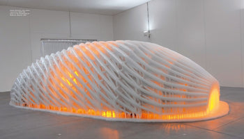 An art installation by Olafur Eliasson in a white gallery, featuring a large, elongated dome sculpture with a lattice framework. The entire structure is covered in a thick layer of white ice and glows intensely from within with a warm, orange light, creating a strong contrast between fire and ice.