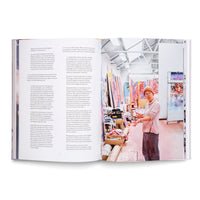 An open book on a white surface, displaying a two-page spread. The left page features text of an interview in German and English. The right page is a full-page photograph of an artist with a beanie and glasses, standing and gesturing in their bright, cluttered studio, surrounded by colorful canvases.