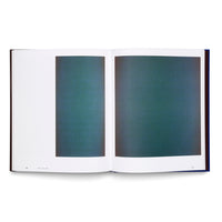 A high-angle photograph of an open art book with a dark blue cover on a white surface. The two-page spread displays an abstract piece featuring two vertical rectangles of a grainy, dark teal color. The rectangle on the left page is narrower than the full-bleed one on the right.