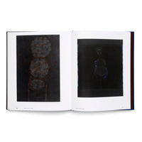An open art book with a dark blue cover, displaying two abstract paintings on a two-page spread. The left page shows a dark painting of three stacked, textured circles in blue and rust. The right page features a minimalist painting of a figure outlined in blue against a black background.