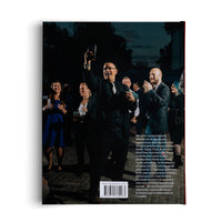 The back cover of a book, featuring a large color photograph of a celebration at night. In the center, a man in a dark suit and glasses joyfully raises a glass in a toast, surrounded by smiling and clapping guests. Below the photo is a block of text in French, a website address, and a barcode.