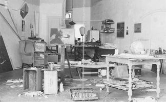 A black and white photograph of a cluttered artist's studio. An easel with a painting stands in the center, surrounded by wooden crates, paint supplies, and papers strewn across the floor. In the background, there is a wingback chair, tables covered in objects, and artwork hanging on the walls.