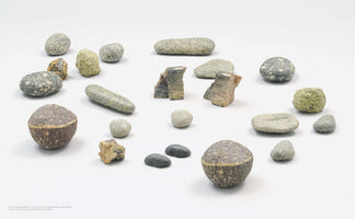 An eye-level studio photograph of various natural stones and sculpted objects arranged on a white surface. The collection includes smooth, speckled river rocks, angular fragments, and two prominent spherical objects with a brown base and a speckled grey top.
