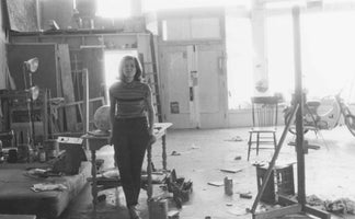 A black and white photograph of a woman with shoulder-length hair standing in a cluttered artist's studio. She wears a striped top and dark pants. The large, brightly lit room is filled with canvases, shelves of supplies, an easel, a wooden table, and a moped in the background.