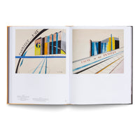 An open art book is displayed against a white background. The two-page spread features colorful, geometric, modernist paintings of abstract factory buildings. The left painting is titled Fabrique de Joie and includes the number 6. The right painting is titled Usine de Mes Pensées. Both artworks use a palette of yellow, blue, orange, and black with strong lines and angular shapes.