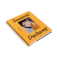 An angled, overhead shot of a hardcover book with a mustard-yellow cover on a white background. The title, Suzanne Duchamp, is printed in a large, stylized black font. In the center is an abstract painting featuring a circular, flower-like geometric object. The spine is visible with the title and names of art institutions.