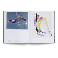 An open art book viewed from above, displaying two colorful abstract paintings. The artwork on the left features geometric shapes and zigzag lines on a blue background. The artwork on the right shows a large, curved, sail-like shape with radiating lines on a white background.